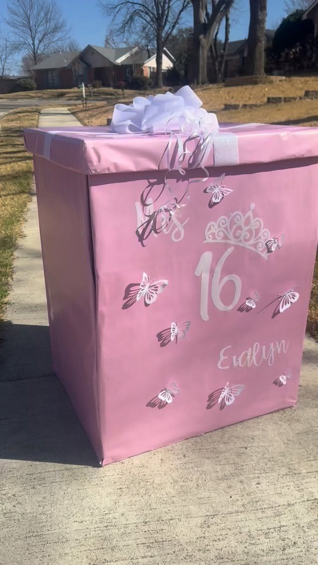Large pink wrapped gift box with white butterflies and number 16 on sidewalk