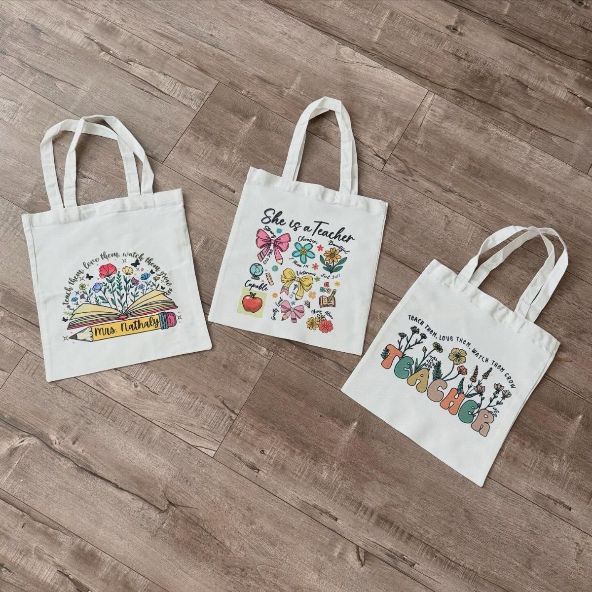 Three white canvas tote bags with teacher-themed colorful prints on a wooden floor.