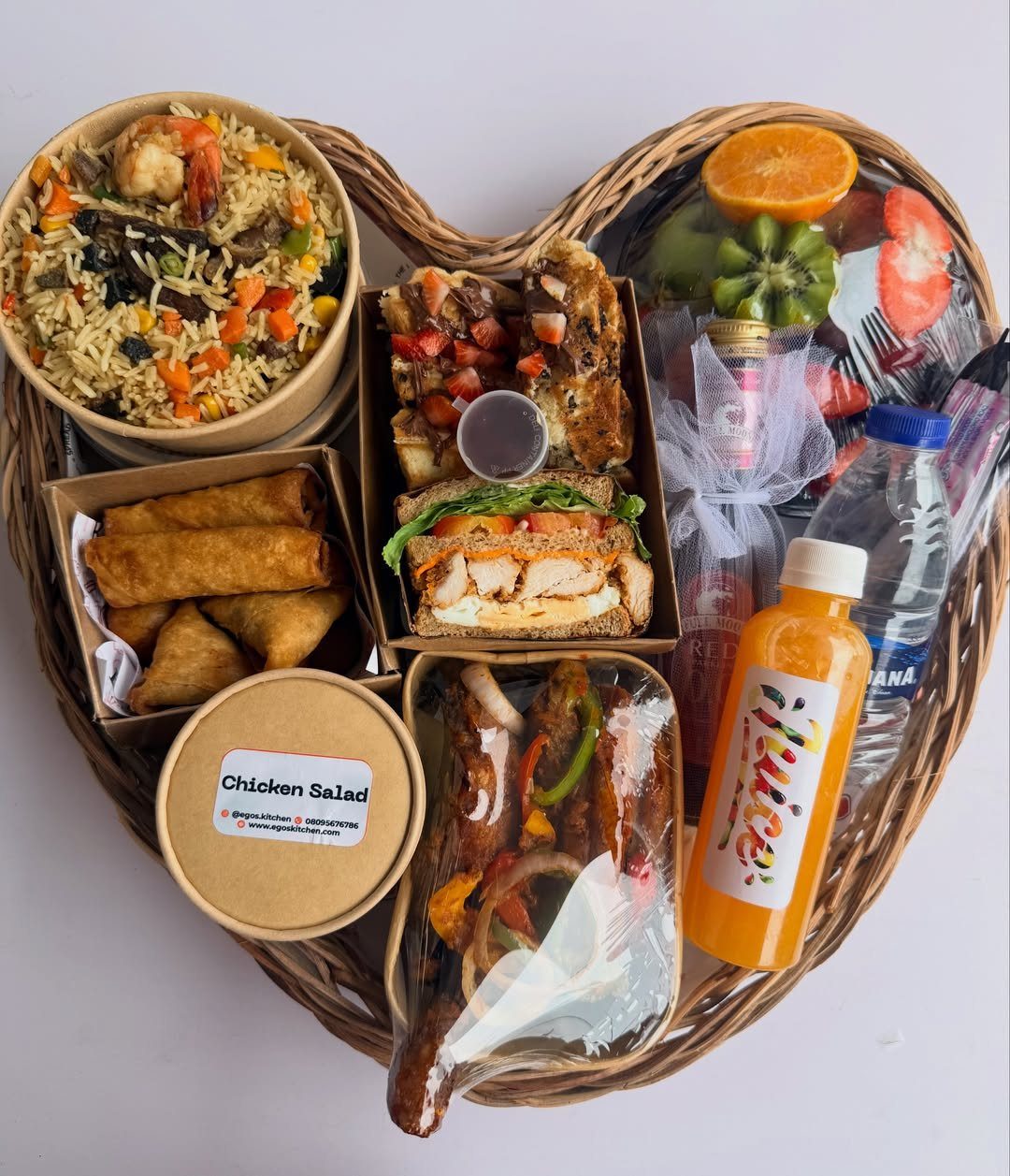 Heart-shaped wicker basket filled with fried rice, sandwiches, grilled meats, and fresh juice.