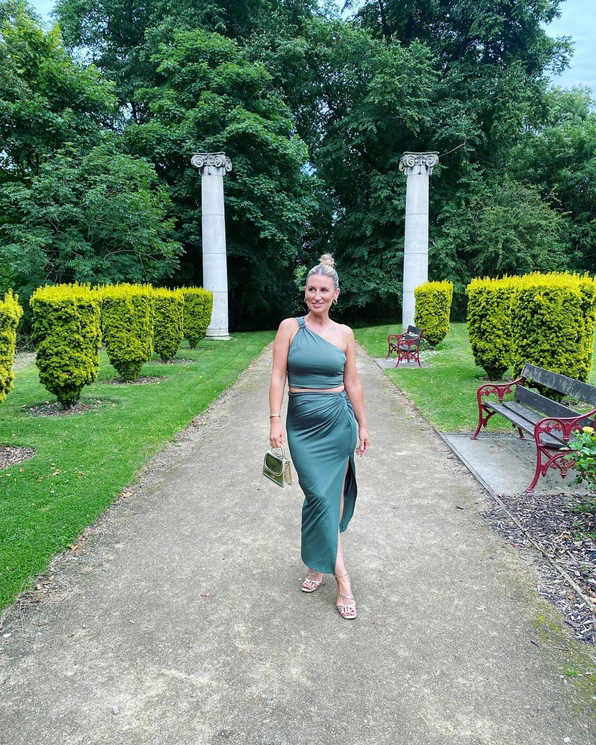 Woman in a green one-shoulder dress holding a small gold clutch in a garden