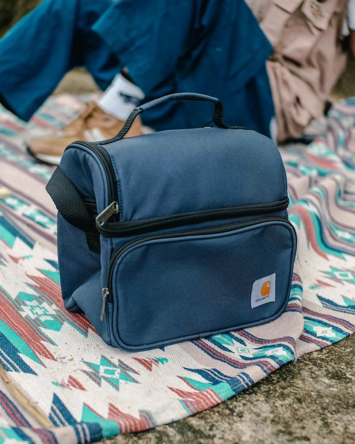 Navy blue Carhartt cooler lunchbox resting on a colorful patterned outdoor blanket.
