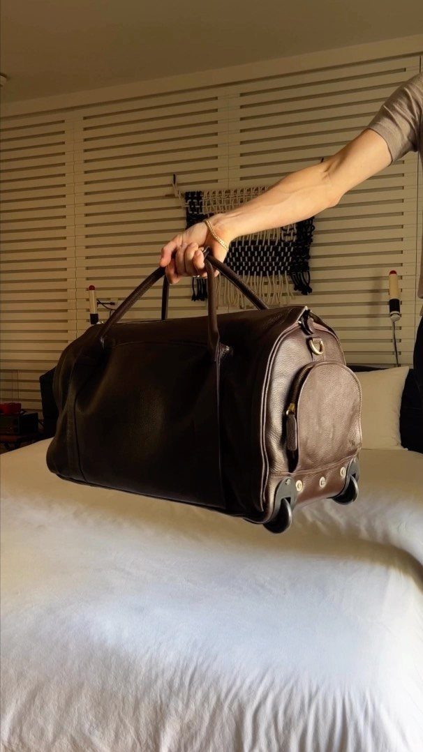 Dark brown leather duffle bag held by a hand over a white bed.