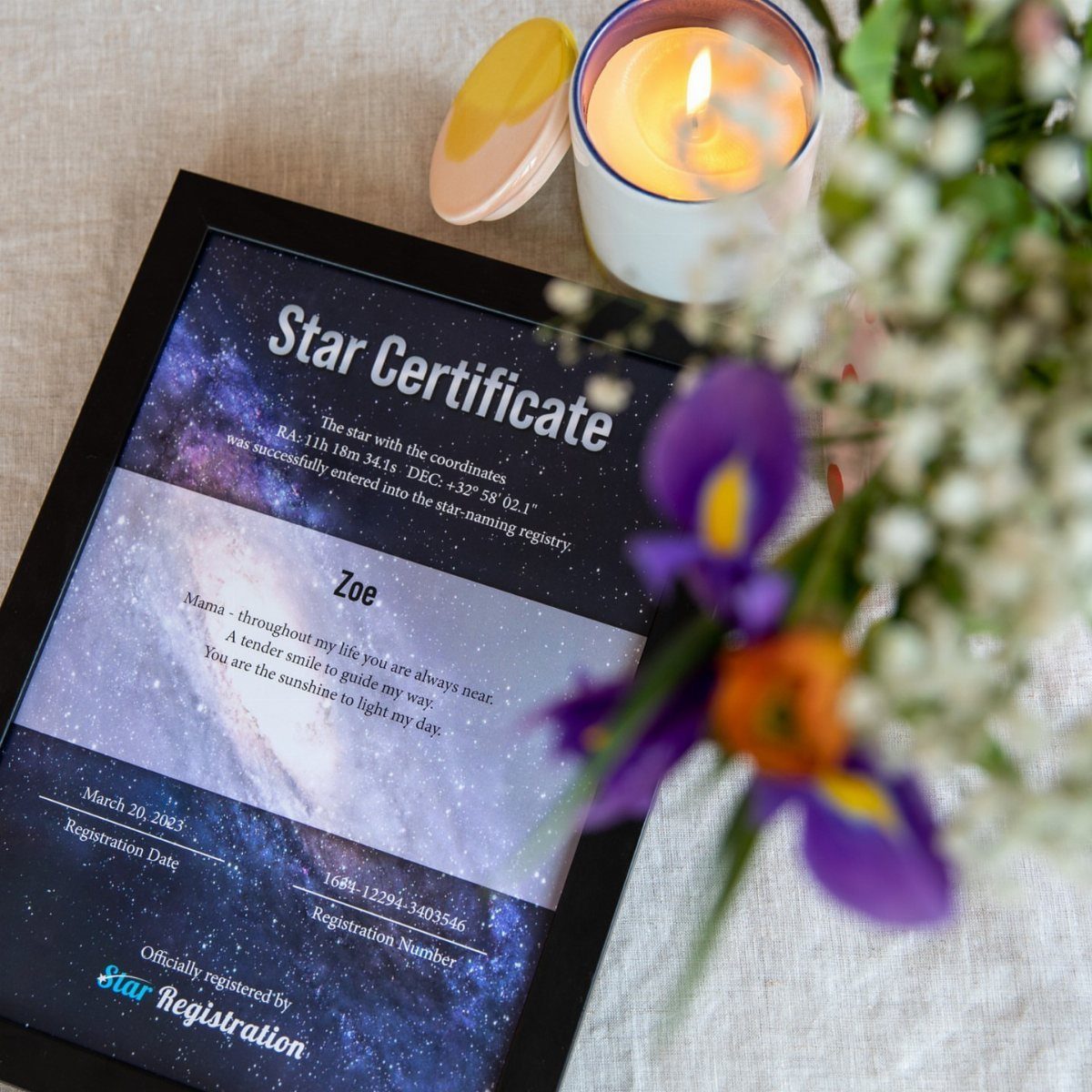 Framed star naming certificate with cosmic imagery next to a candle and flowers.