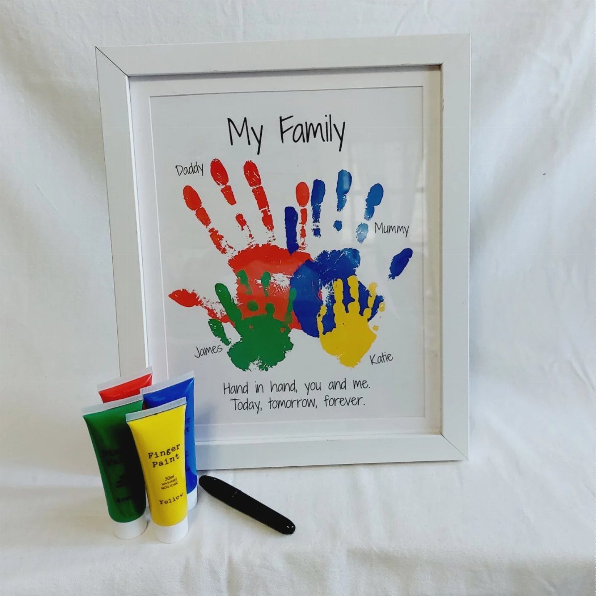 Framed family handprint art with colorful paint tubes and a marker in front.
