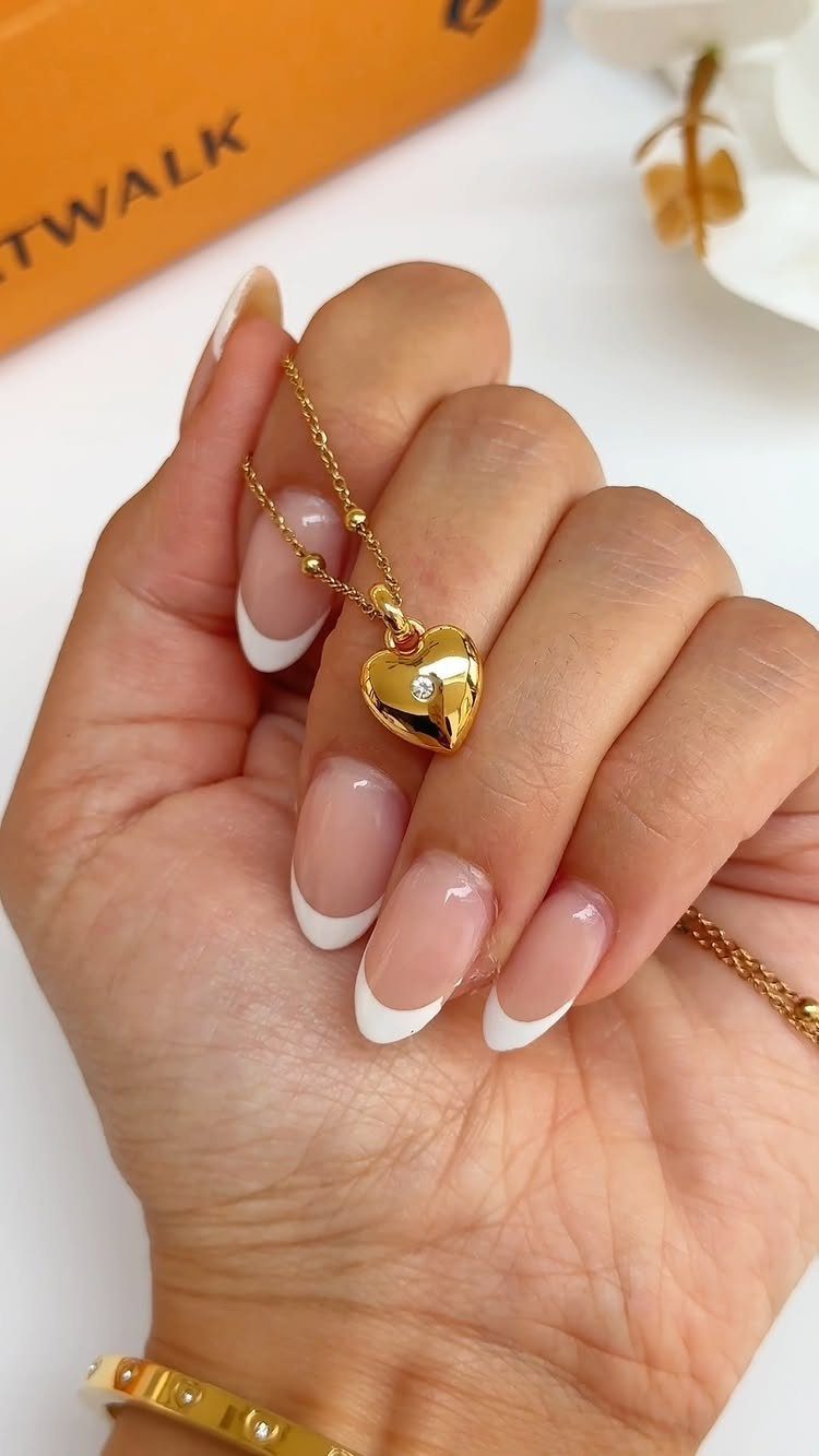 Hand with French manicure holding a gold heart pendant necklace and wearing a bracelet.