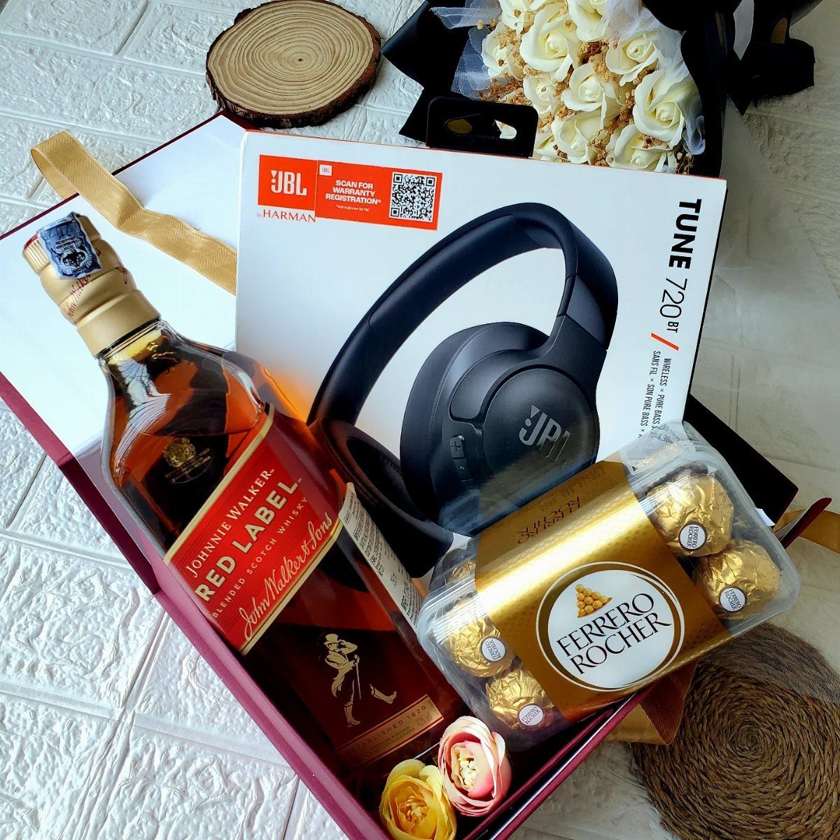 Gift box with whiskey bottle, black headphones, Ferrero Rocher chocolates, and small roses.