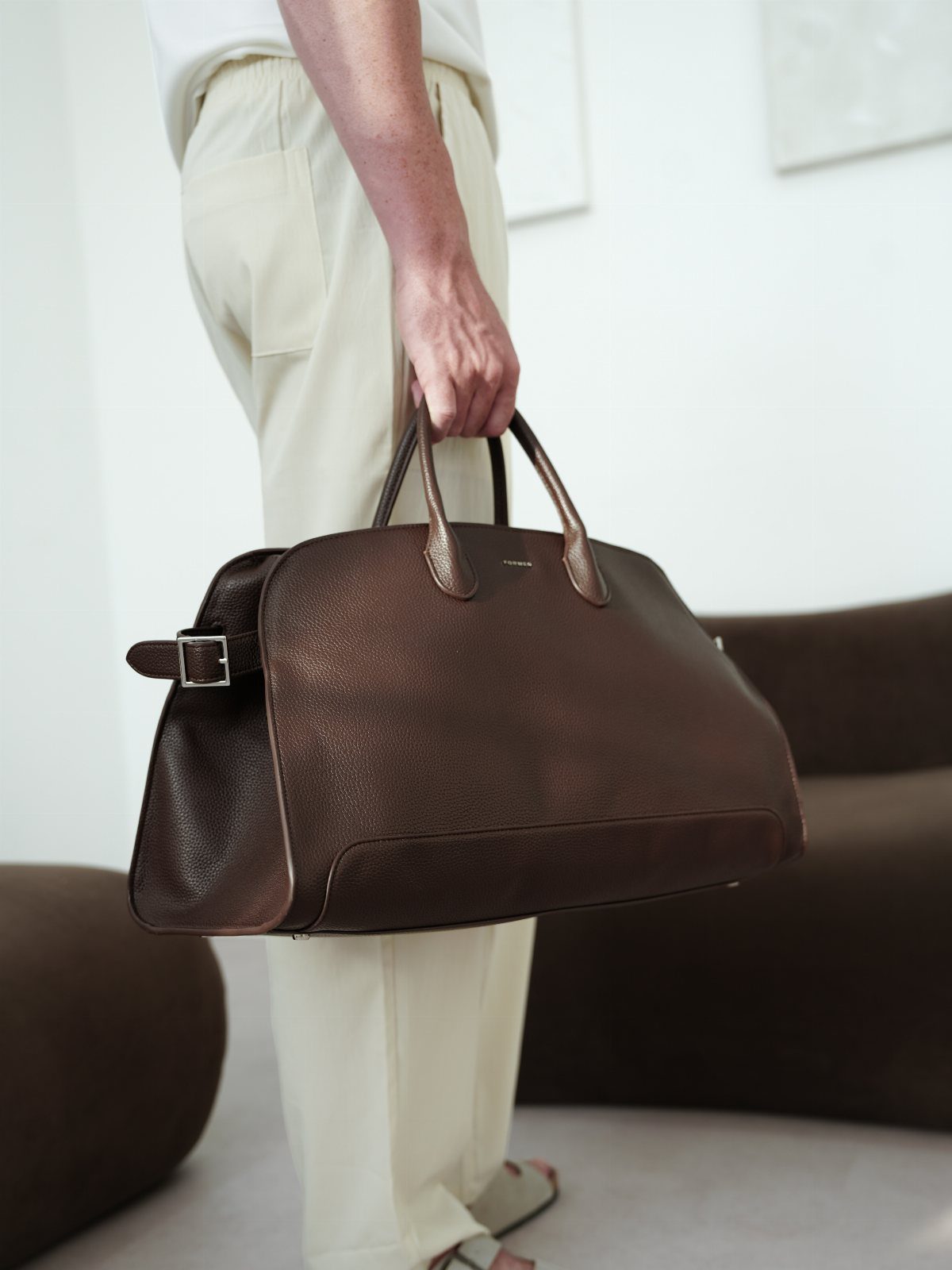 Dark brown leather duffel bag held by a person in cream trousers.