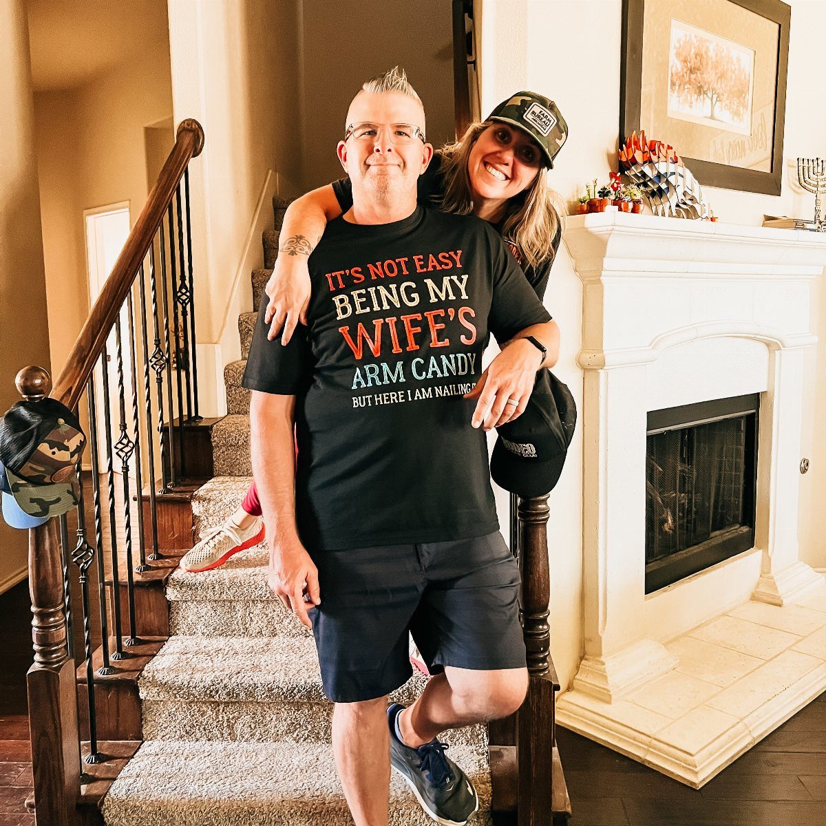 Man wearing a funny husband t-shirt while being hugged by his wife indoors.
