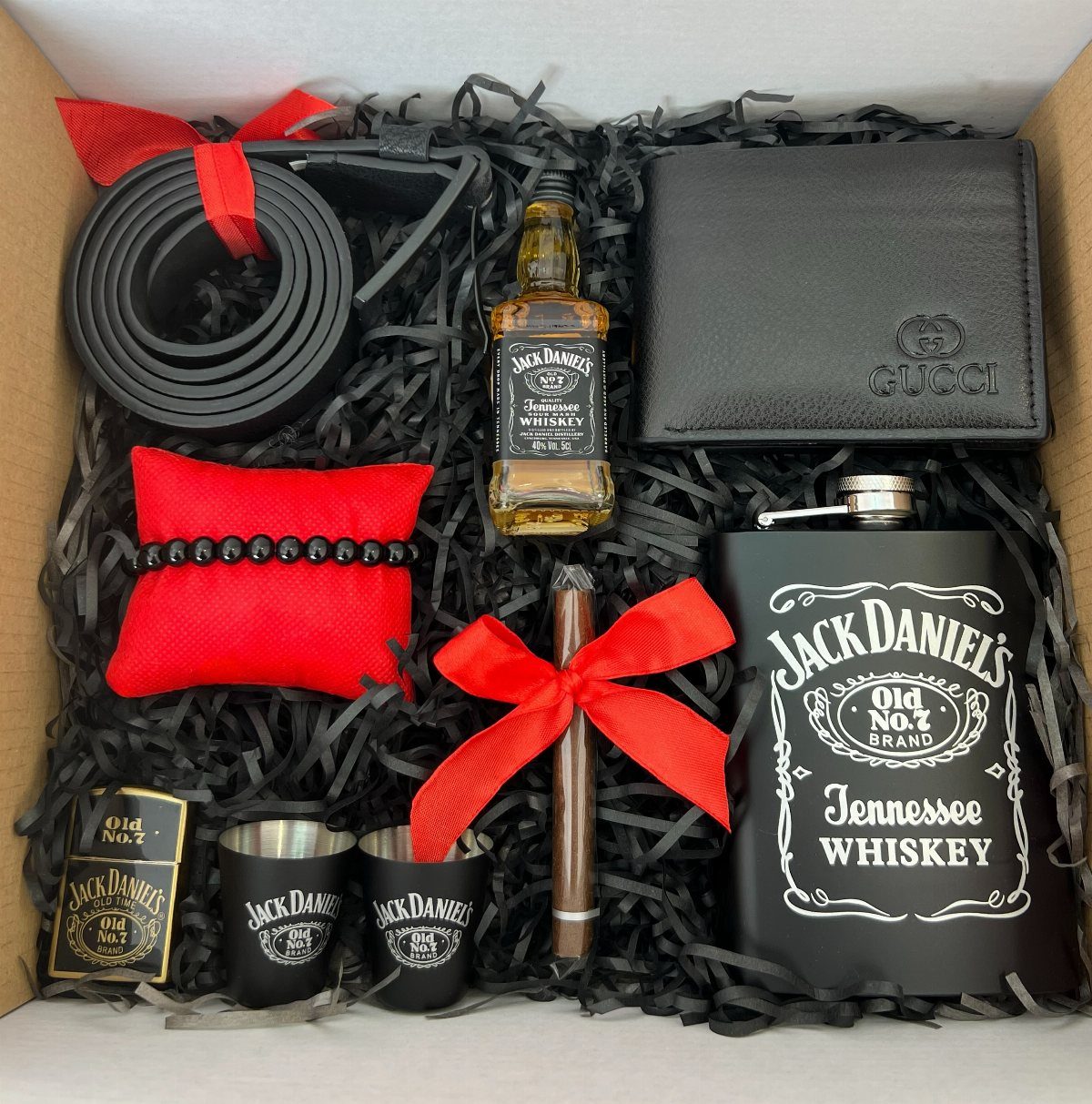 Gift box with whiskey bottle, flask, cigar, black wallet, and belt on black filler.