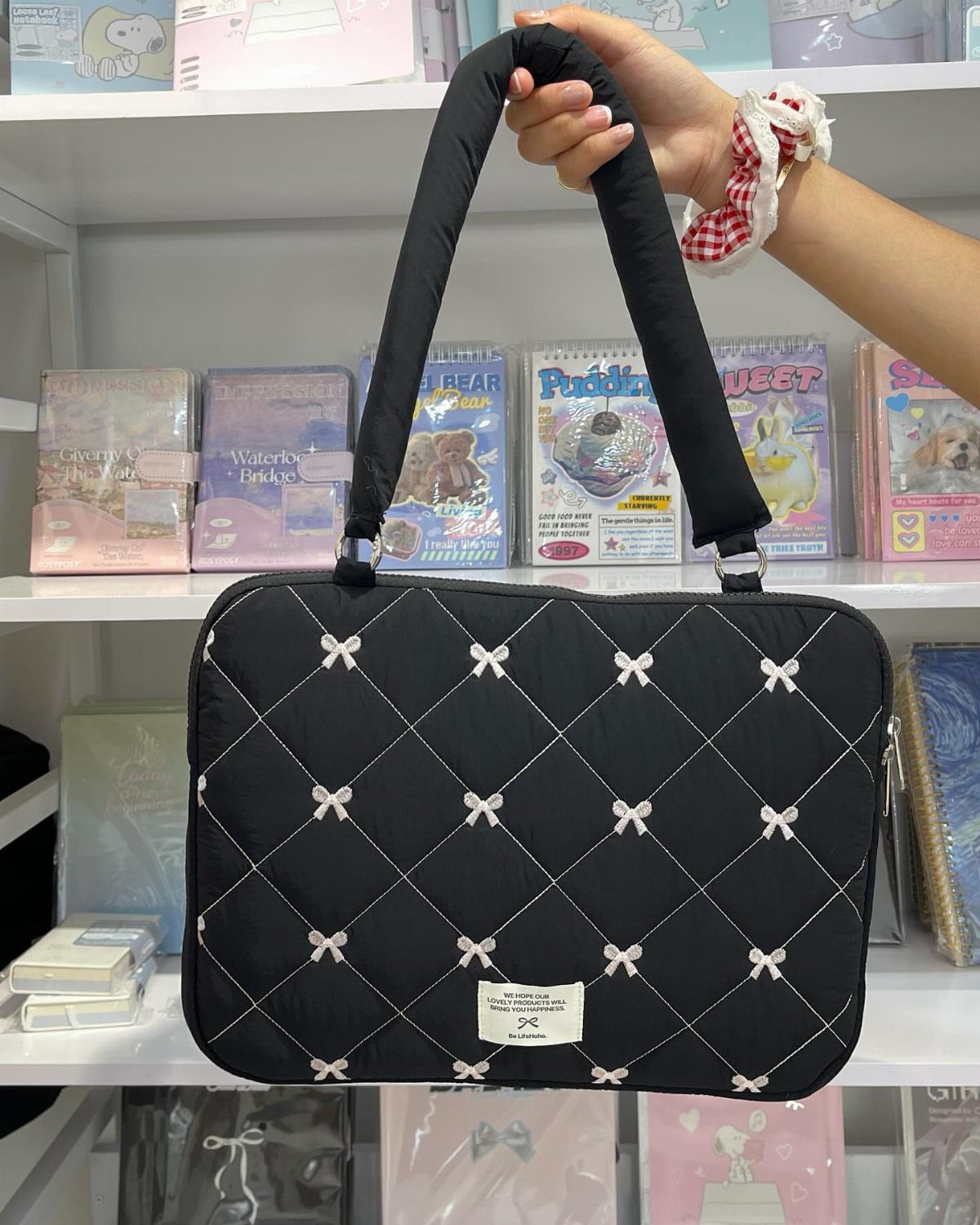 Black quilted laptop bag with white bows held in front of stationery shelves