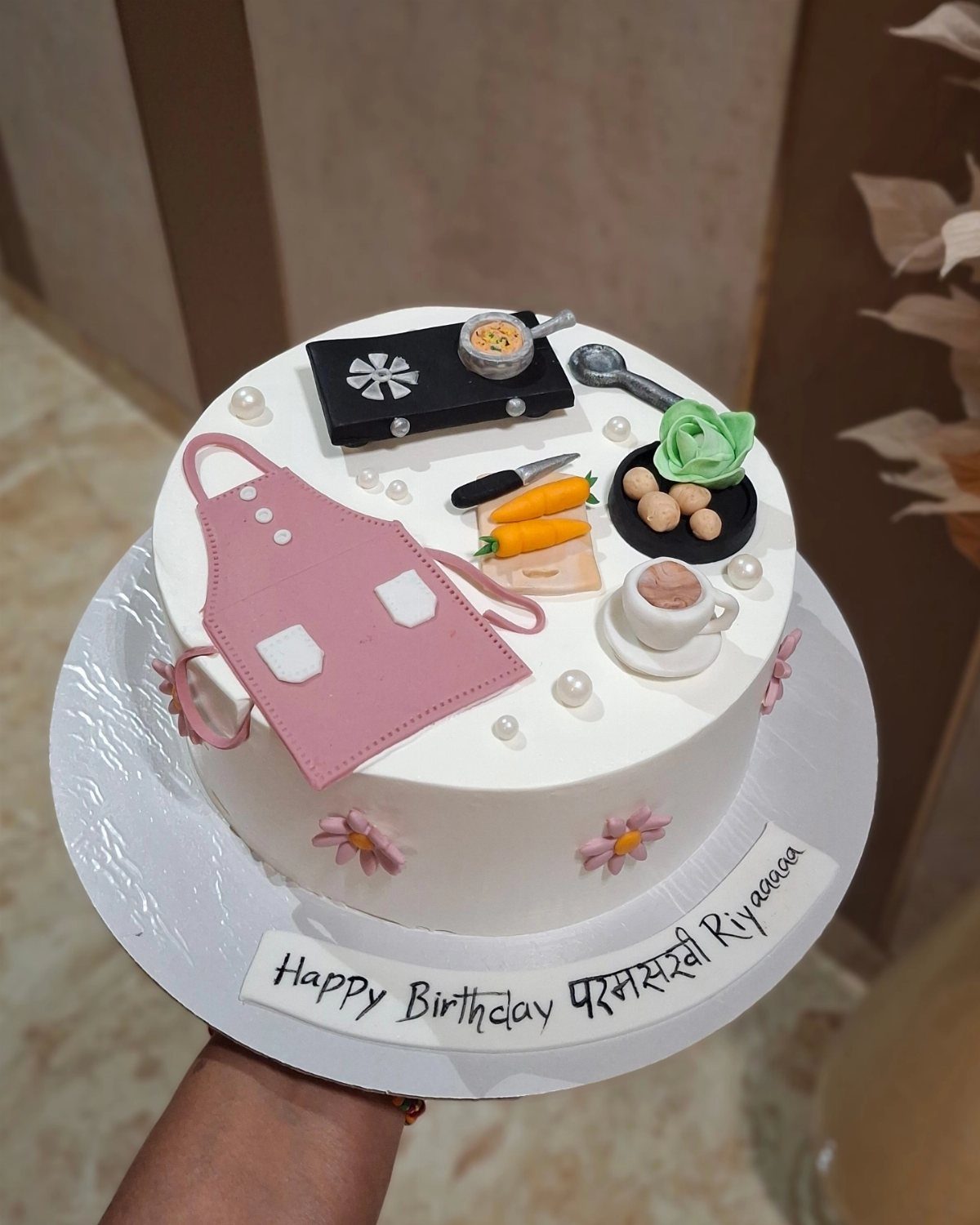 White birthday cake decorated with fondant cooking tools and a pink apron.