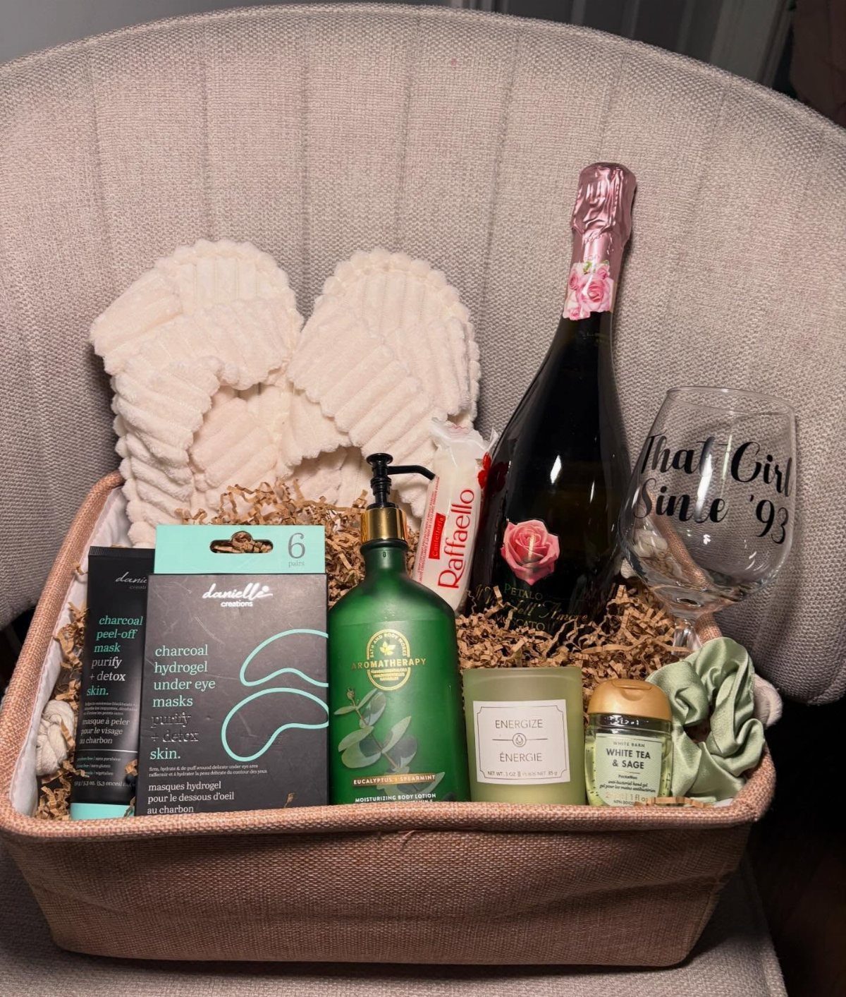 Gift basket with champagne, skincare products, a candle, and fluffy white slippers.