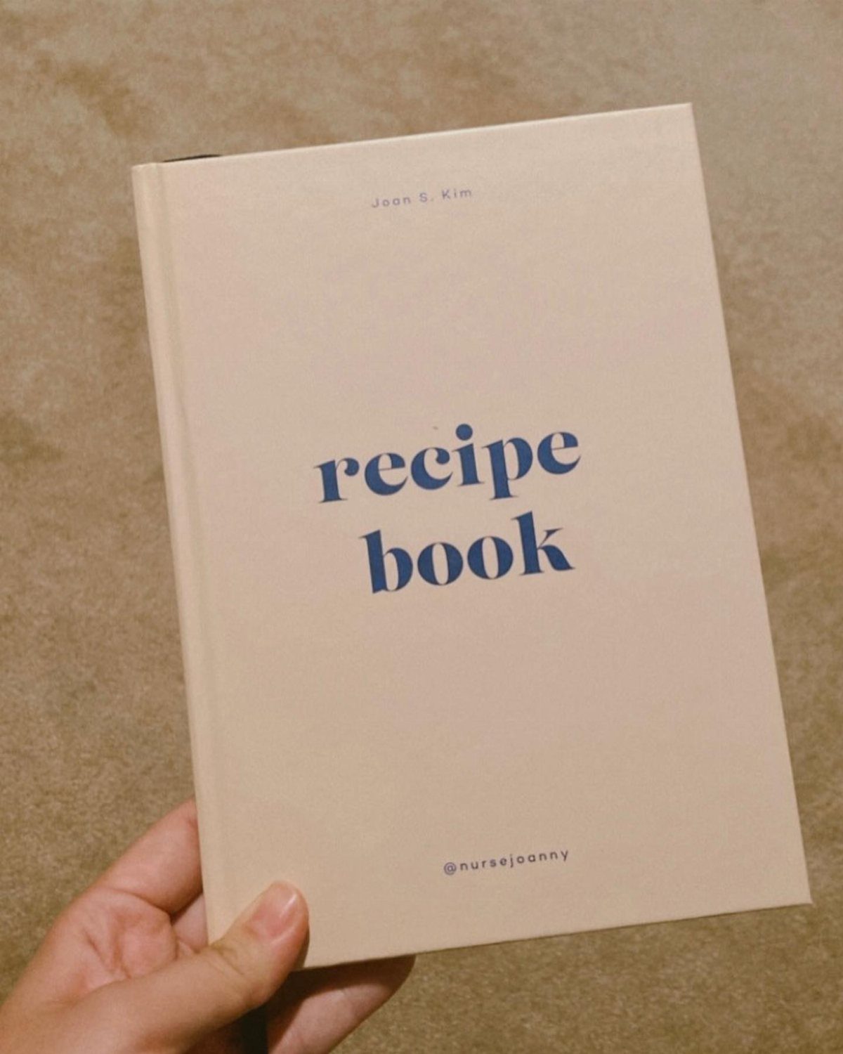 Cream hardcover recipe book with blue text held by a person's hand.