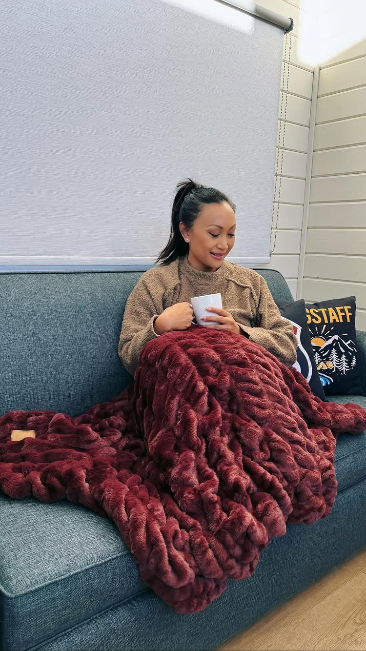 Woman wrapped in a thick burgundy faux-fur blanket sitting on a grey sofa