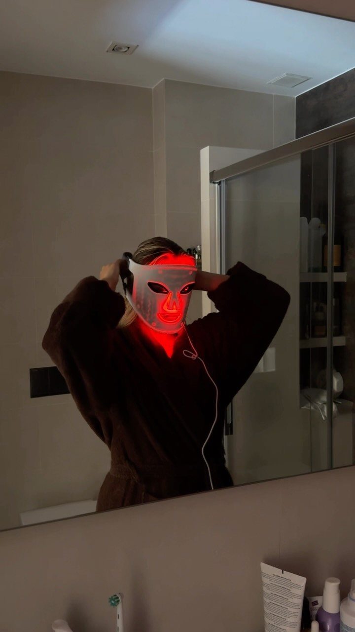 Person in a brown robe wearing a glowing red light therapy face mask.