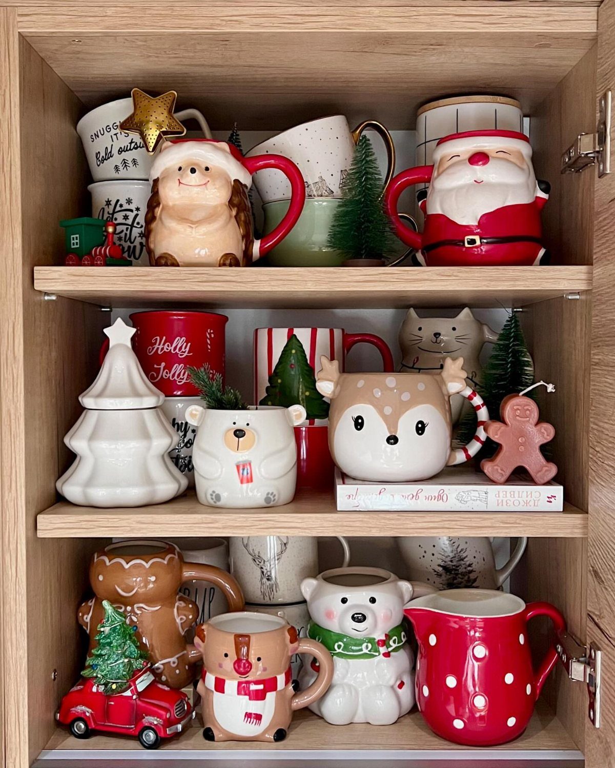 Festive ceramic mugs and Christmas figurines arranged on wooden cabinet shelves.