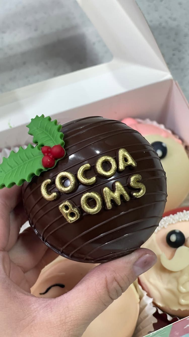 Chocolate cocoa bomb with gold lettering and holly decoration held in a hand.